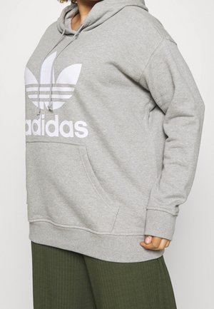 Osoba ubrana w szarą bluzę z kapturem Adidas z białym logo oraz oliwkowozielone prążkowane spodnie, stojąca na jednolitym tle.