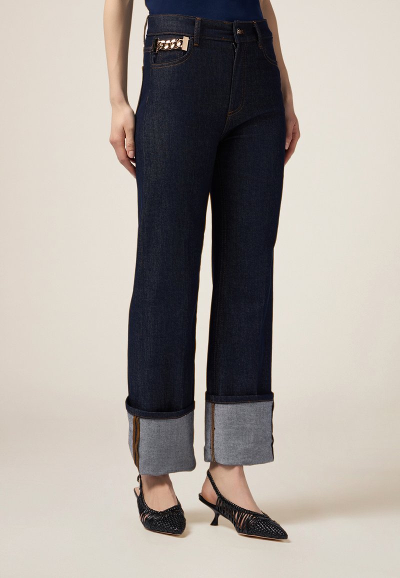 Jeans in denim scuro a gamba larga con orli risvoltati in tessuto blu chiaro. Caratterizzati da un dettaglio in catena dorata sulla tasca frontale.
