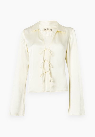 Blouse crème à manches longues avec un col et un décolleté en V, présentant un devant à lacets avec trois liens en tissu et une texture lisse et soyeuse.
