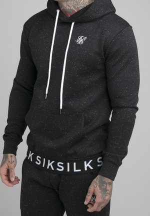 Mężczyzna ma na sobie czarną bluzę z kapturem w jasne plamki z białymi troczkami, kieszenią, logo SikSilk na piersi oraz dopasowane spodnie z paskiem SikSilk; widoczne tatuaże na rękach.