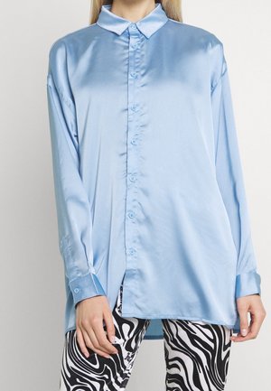 Vaaleansininen satiininen pitkähihainen nappipaita ja mustavalkoiset abstraktikuvioiset housut päällä seisovalla henkilöllä.