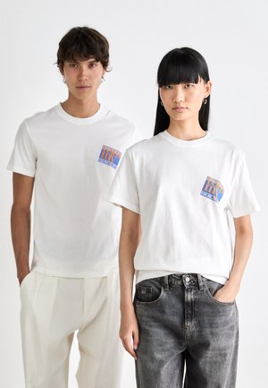 Ung mand og kvinde iført hvide Tommy Jeans t-shirts med logoer, stående side om side mod en simpel hvid baggrund.