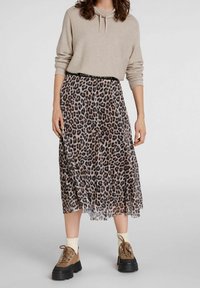 Hame pleated kuosisimmon kanssa, jossa on läpinäkyvä päällinen, yhdistettynä beigeen huppariin ja ruskeisiin paksupohjiin kenkiin. Vyötäröllä on musta brändin elastinen nauha.