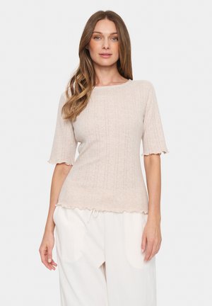 Maglione in maglia color beige chiaro con maniche corte e ondulate. Presenta una texture a righe verticali e un orlo scallop per un rifinitura morbida. Abbinato a pantaloni bianchi.