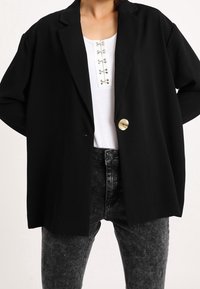 Person med en svart oversized kavaj med en guldknapp, vit skjorta med silverkläppen och svarta acid-wash jeans mot en vit bakgrund.