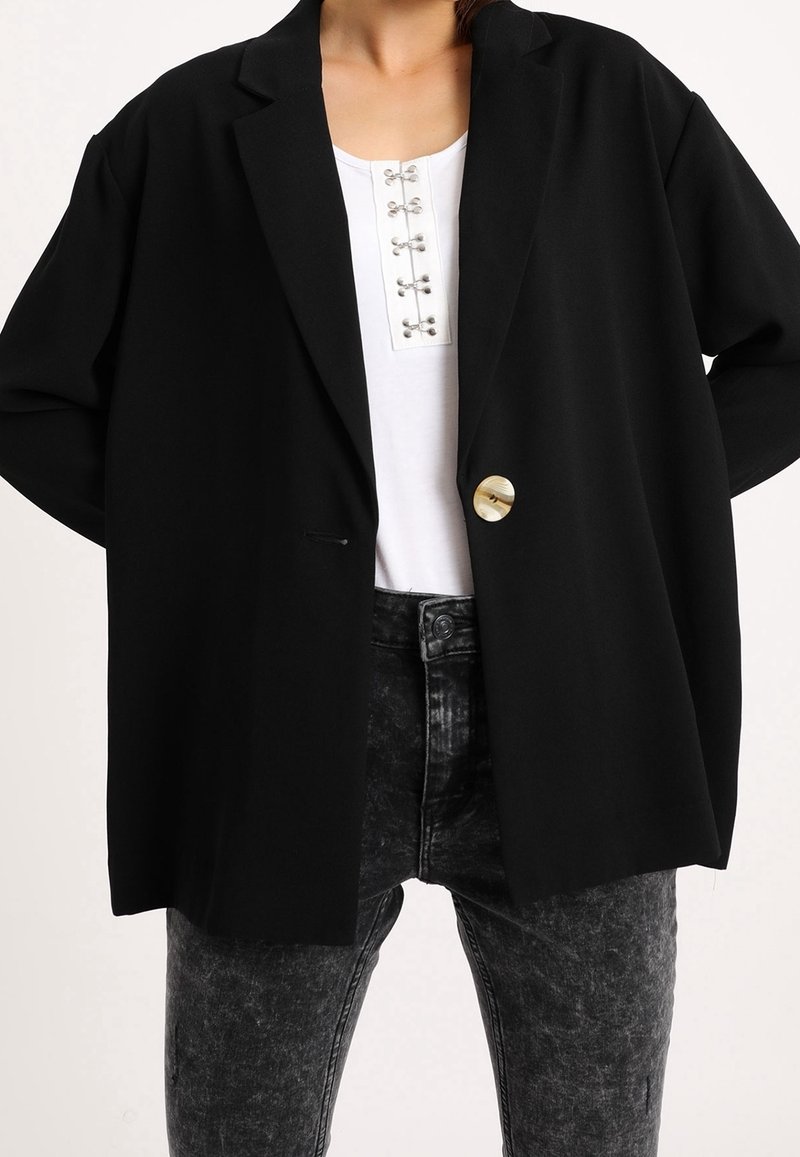 Person med en svart oversized kavaj med en guldknapp, vit skjorta med silverkläppen och svarta acid-wash jeans mot en vit bakgrund.