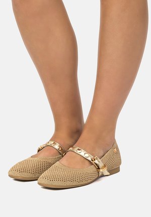 Pies con zapatos planos tejidos beige con correas tachonadas de oro y detalles de hebilla dorada sobre un fondo claro.