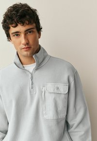 Lysegrå pullover-sweatshirt lavet af blødt stof, med lynlåslukning ved kraven og en brystlomme med en knap. Tekstureret finish.