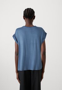 Haut bleu à manches courtes avec une coupe décontractée, une texture lisse et des poignets retroussés, présenté de dos sur un fond clair.