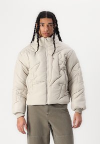 Ljusbeige puffer-jacka med hög krage, vadderad textur och abstrakt mönster. Har en dragkedja framtill och ribbade muddar.