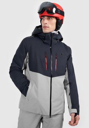 Man in black and gray ski jacket and pants wearing red and black ski helmet, looking to the side with one hand in jacket pocket.