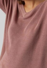 Gros plan sur une personne portant un pull en maille rose poussiéreux à col en V, montrant la texture et la coupe du tissu sur le haut du torse.