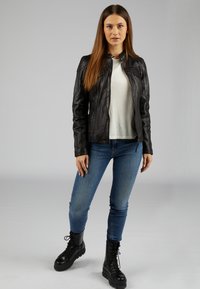 Schwarze Lederjacke mit Reißverschluss, taillierte Passform. Kombiniert mit einem hellen Hemd und blauen Jeans, abgerundet mit schwarzen Schnürkampfschuhen.