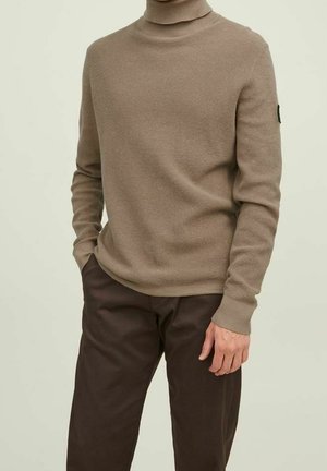 Suéter de cuello alto acanalado beige con mangas largas, hecho de un material de punto texturizado; combinado con pantalones marrón oscuro. Acento negro del logo en la manga.
