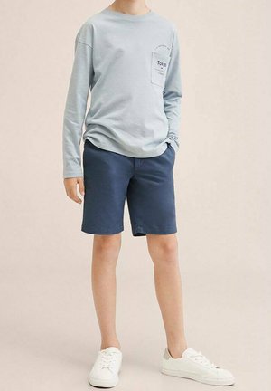 Barn iført en lysegrå langærmet trøje med teksten "Tokyo" på lommen, navy blå shorts og hvide sneakers står med den ene hånd i lommen.
