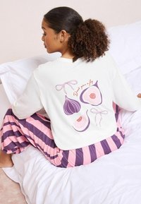 Fehér pulóver fügeillusztrációkkal és "Don't give a fig" felirattal a hátsó részén; lila és rózsaszín csíkozott pizsamabugyival párosítva.