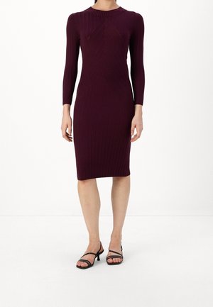 Woman wearing a fitted, ribbed, long-sleeve, knee-length dark purple dress with black strappy low-heeled sandals on a white background.