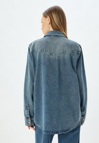 Camisa de mezclilla en azul claro con mangas largas, puños abotonados, cuello puntiagudo y un dobladillo curvado; presenta un sutil detalle de costura en la parte posterior.