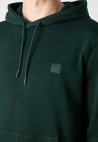 Sweat à capuche vert foncé avec cordons et petite écusson carré portant la mention « BOSS » sur la poitrine gauche, porté par une personne.