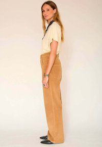 Haut beige à manches courtes, pantalon en velours côtelé marron taille haute à jambes larges, et chaussures noires à bout pointu avec des clous en argent. Des accessoires simples complètent le look.