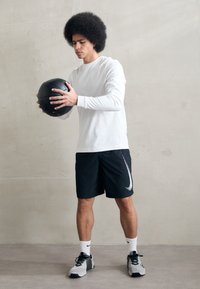 Un ballon de médecine noir tenu par une personne portant une chemise blanche à manches longues et un short noir avec le swoosh blanc de Nike. Fond gris.