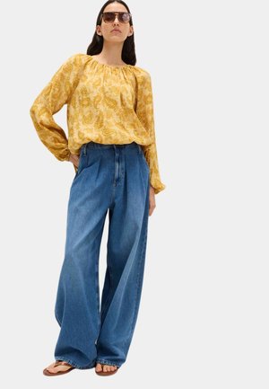 Femme portant un jean bleu large oversize, un chemisier jaune à manches longues à motifs, des lunettes de soleil et des sandales marron, debout avec une main sur la hanche.