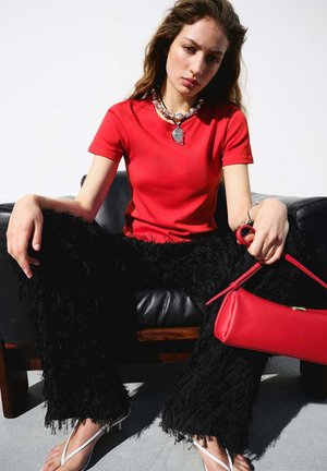 Mujer joven con camiseta roja, pantalones negros con textura, tacones blancos y collar plateado, sentada en una silla de cuero negra sosteniendo un bolso rojo.
