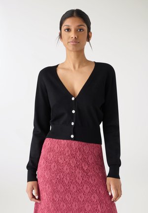 Czarny dopasowany kardigan z głębokim dekoltem w kształcie litery V, zdobiony pięcioma guzikami w kształcie kwiatów. Noszony na teksturowanej różowej koronkowej spódnicy.