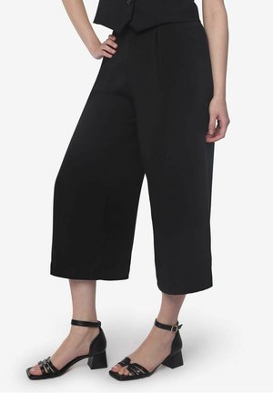 Culottes noires en tissu léger, présentant une coupe à jambes larges tombant sous le genou, assorties à des talons noirs à brides.