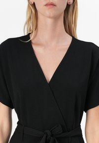 Černé zavinovací šaty s krátkými rukávy, výstřihem do V a uvázaným páskem v pase. Vyrobeno z hladké tkaniny s čistým a minimalistickým designem.
