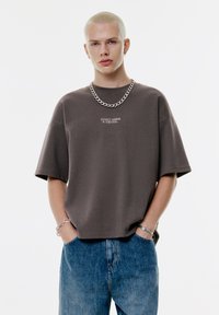 Ung person med platinablond hår bærer en kæderekreation, oversized mørkegrå T-shirt med teksten "GÅ IKKE GLIP AF EN DETALJE" og blå jeans, hænderne i lommen.