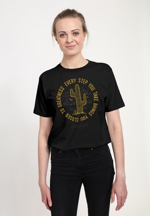 Kvinde iført sort t-shirt med gul kaktusgrafik og teksten "Hvert skridt du tager, bringer dig nærmere storhed" på forsiden.