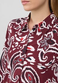 Close-up van een persoon die een knoopshirt in kastanjebruin draagt met witte, grijze en rode bloemen- en paisleypatronen.