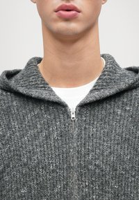 Sivá pletená mikina na zips s textúrovaným vzorom, s kapucňou a kovovým zipsom. Pod ňou je biele tričko s okrúhlym výstrihom.