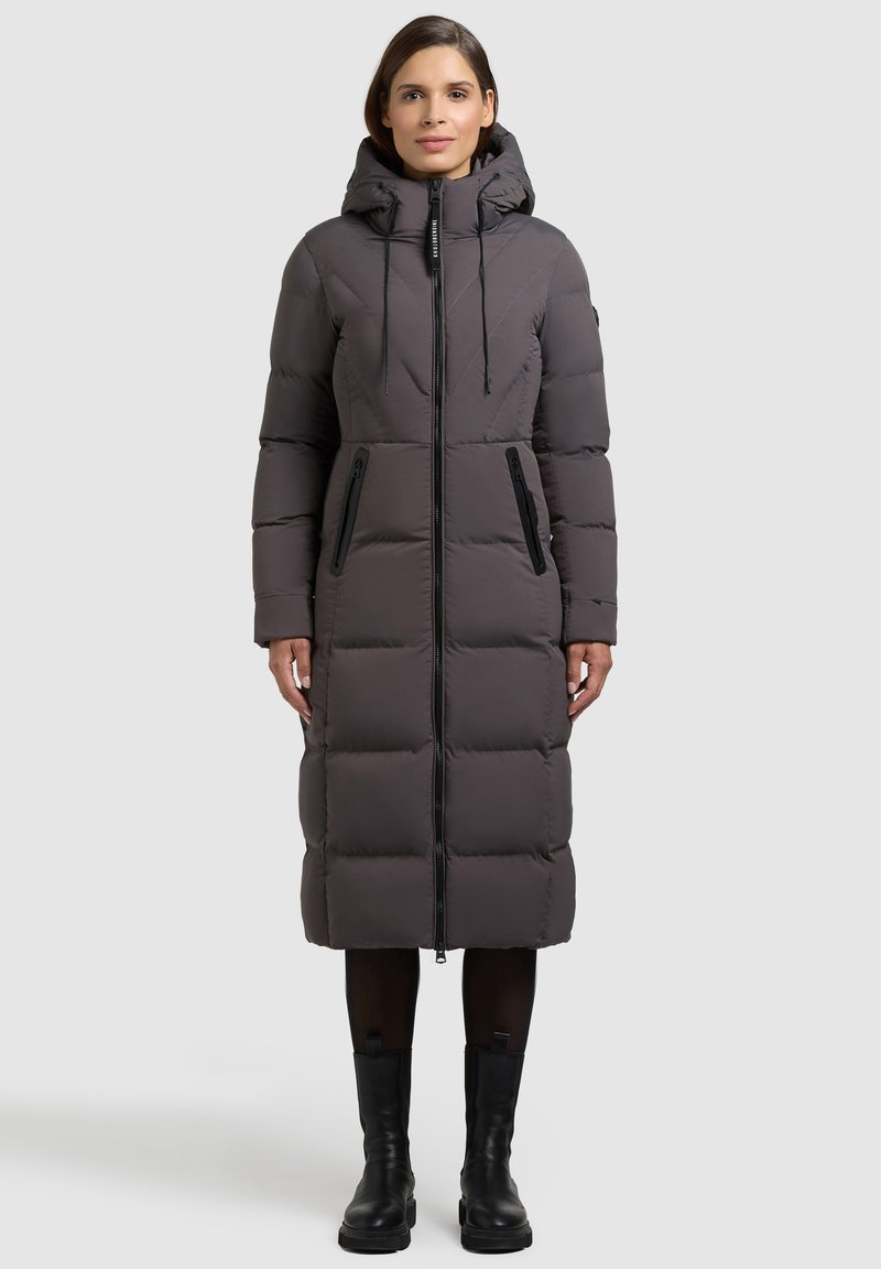 Pitkä, tikattu toppatakki tummanharmaana, jossa on huppu, edessä vetoketjusuljenta ja kaksi vetoketjullista sivutaskua, yhdistetty mustiin saappaisiin.