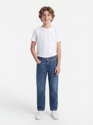 Teenager-Junge mit lockigem Haar steht lächelnd mit einer Hand in der Tasche, trägt ein weißes T-Shirt, blaue Jeans und weiße Turnschuhe vor weißem Hintergrund.