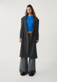 Cappotto doppio petto in misto lana grigio con vestibilità over, abbinato a un crop top blu aderente e pantaloni grigi a gamba larga. Scarpe platform nere.
