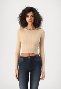 Crop top côtelé beige clair avec manches longues, présentant un col rond. Associé à un jean ajusté bleu foncé.