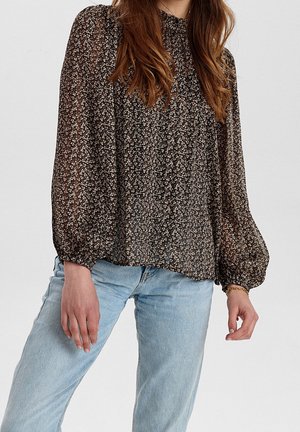 Vrouw draagt een zwarte bloemenlange mouwen blouse met pofmouwen en lichtblauwe spijkerbroek tegen een effen achtergrond.