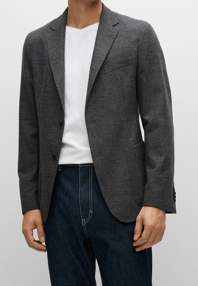 Blazer ajusté gris foncé en tissu texturé, doté de deux boutons, de revers crantés et de poches latérales. Porté sur un t-shirt blanc et un jean foncé.