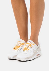 Baskets Nike avec une tige en cuir blanc et mesh, des accents en daim gris, des lacets orange et un amorti visible dans la semelle. Semelle extérieure noire.