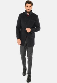 Cappotto in misto lana di colore blu navy con colletto alto, chiusura frontale a bottoni, tasche laterali e vestibilità sartoriale. Abbinato a pantaloni grigi e scarpe nere.