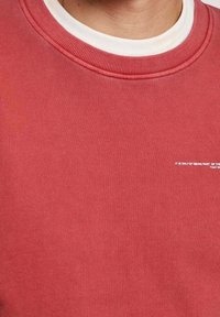 Rotes Sweatshirt mit einem gerippten Rundhalsausschnitt und einer weißen inneren Kragenlage. Enthält subtile Textdetails auf der Vorderseite. Glatte Textur.