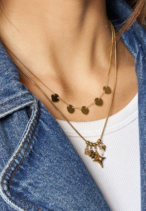 Gelaagde gouden kettingen: de ene heeft ronde hangers, de andere heeft bedels waaronder een ster en een hart, tentoongesteld tegen een spijkerjacket.
