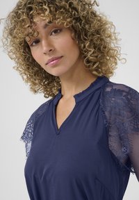 Navy blouse with short lace sleeves, V-neck design, and ruffled collar. Fabric has a smooth texture, detailed with delicate lace patterns.
