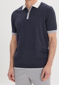 Granatowa koszulka polo z krótkim rękawem z białym kołnierzykiem, szarymi akcentami przy rękawach i plisą. Wykonana z gładkiego materiału, dopasowany krój.