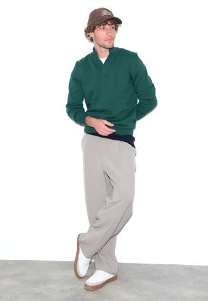 Homme portant une casquette marron, un pull vert foncé, un pantalon beige et des baskets blanches, debout, jambes croisées, regardant sur le côté.