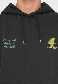 Hoodie noir en tissu mélangé de coton. Dispose d'une poche avant, d'une capuche avec cordon de serrage et du texte : "Vêtements de Sport Universels" et "4ème quart" en bleu et jaune.
