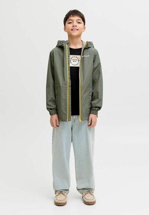 Dreng stående og smilende, iført en olivengrøn jakke med gul lynlås, sort T-shirt med grafik, lyseblå jeans og beige sko.