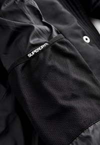 Poche en tissu mesh noir étiquetée "SUPERDRY" sur une veste noire lisse avec un bouton pression argenté visible.